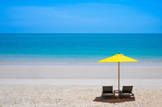 Tropical summer sand beach with two lounge chairs under a yellow umbrella against blue sea background in sunny day. Idyllic travel and summer vacation concept.
