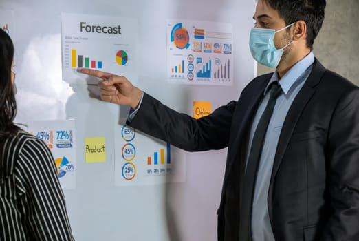 Young businessman with protective face mask pointing towards graph while giving presentation in office. Business marketing information concept.