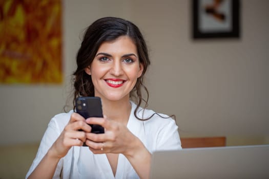 Businesswoman smiling to the camera from home office. Happy female emplyee working from home with positive mindset. Satisfied young adult feminine holding mobile in hands.