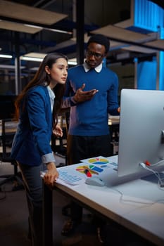Man and woman looks on screen, office lifestyle. Male and female persons talking at laptop, dark interior on background, modern workplace