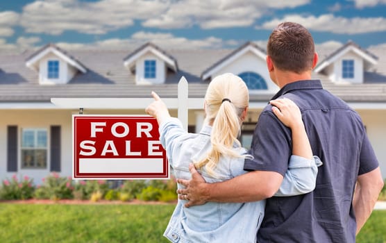 Young Adult Couple Facing and Pointing to Front of For Sale Real Estate Sign and House.