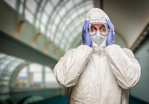 Man Holding Head With Hands Wearing HAZMAT Protective Clothing Inside Building.