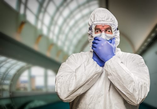 Man Covering Mouth With Hands Wearing HAZMAT Protective Clothing Inside Building.
