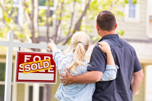 Caucasian Couple Facing and Pointing to Front of Sold Real Estate Sign and House.