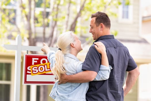Caucasian Couple Facing and Pointing to Front of Sold Real Estate Sign and House.
