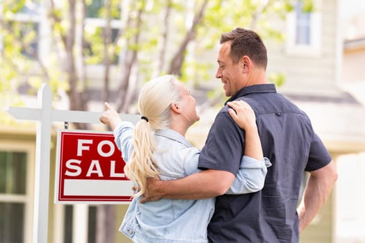 Caucasian Couple Facing and Pointing to Front of For Sale Real Estate Sign and House.
