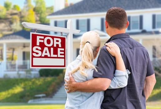 Young Adult Couple Facing Front of For Sale Real Estate Sign and House.