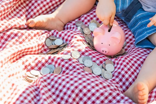 Baby Boy Sitting on Picnic Blanket PUtting Coins in Piggy Bank