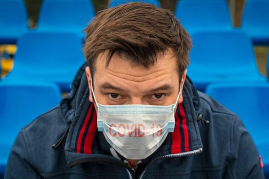 Man in surgical mask sits alone in stadium. COVID-19 Pandemic Coronavirus. Man in city stadium wearing face mask protective for spreading of Coronavirus Disease 2019. Portrait of man with surgical mask on face against virus.