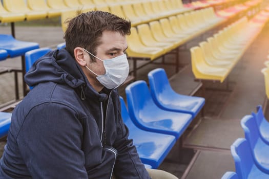 Man in surgical mask sits alone in stadium. COVID-19 Pandemic Coronavirus Man in city stadium wearing face mask protective for spreading of Coronavirus Disease 2019. Portrait of man with surgical mask on face against virus.