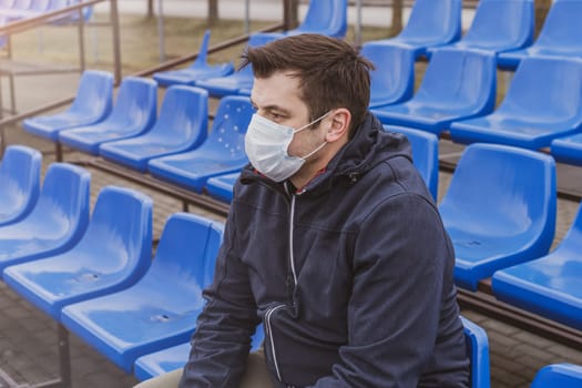 Man in surgical mask sits alone in stadium. COVID-19 Pandemic Coronavirus Man in city stadium wearing face mask protective for spreading of Coronavirus Disease 2019. Portrait of man with surgical mask on face against virus.