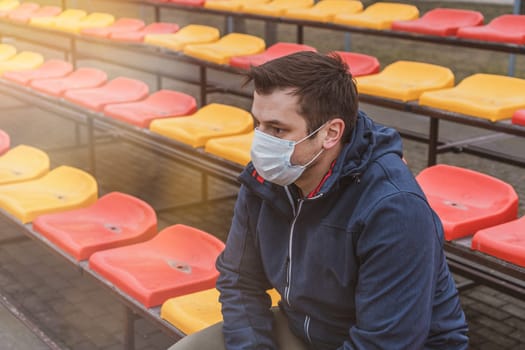 Man in surgical mask sits alone in stadium. COVID-19 Pandemic Coronavirus. Man in city stadium wearing face mask protective for spreading of Coronavirus Disease 2019. Portrait of man with surgical mask on face against virus.