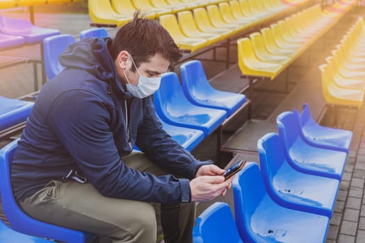 Young man wearing a health mask, while looking at his mobile phone.Man in surgical mask sits alone in stadium. COVID-19 Pandemic Coronavirus. Man in city stadium wearing face mask protective for spreading of Coronavirus Disease 2019.