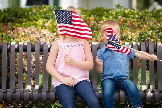Young Sister and Brother Waving American Flags On The Bench At The Park.
