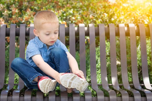 Young Boy Sitting On A Bench Putting On His Shoes At The Park.