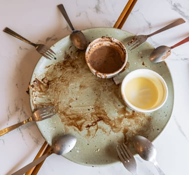Empty plate with food leftovers from dessert meal, spoons and forks on marble table. Dirty plate with chocolate sauce, chocolate and vanilla ice cream leftovers in ramekin bowl.