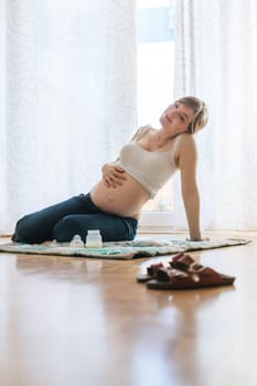 Happy Caucasian pregnant mother is sitting on the floor, touching her tummy, blue jeans