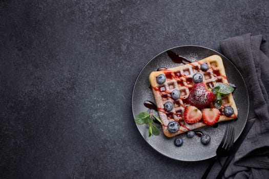 Delicious waffles with berries, mint, powdered sugar and sweet sauce on ceramic black plate on grey background. Top view with copy space. Sweet meal. Dessert. Serving food