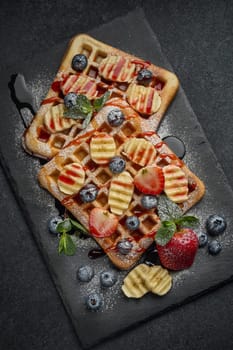 Delicious waffles with berries, mint, powdered sugar and sweet sauce  on slate serving board on grey background. Top view. Sweet meal. Dessert. Serving food