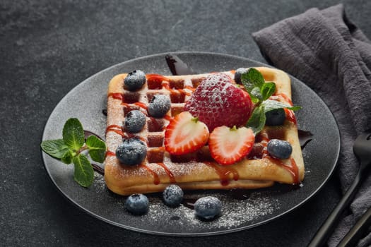 Delicious waffles with  powdered sugar and berries on ceramic black plate on grey background. Sweet meal. Dessert. Serving food