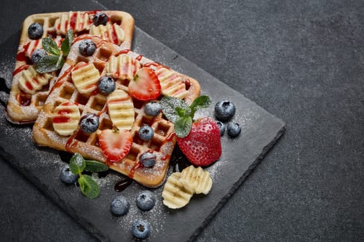 Delicious waffles with berries, mint, powdered sugar and sweet sauce  on slate serving board on grey background. Top view. Sweet meal. Dessert. Serving food