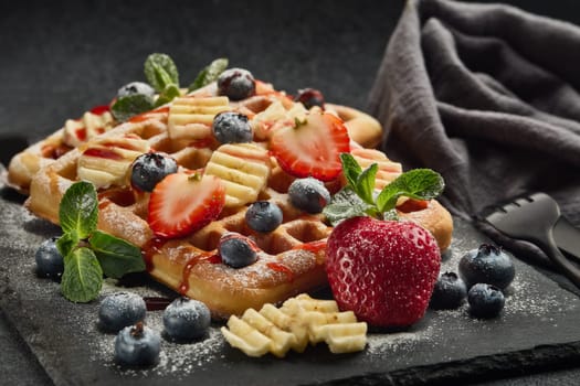 Delicious waffles with berries, mint, powdered sugar and sweet sauce  on slate serving board on grey background. Sweet meal. Dessert. Serving food