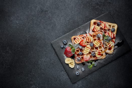 Delicious waffles with berries, mint, powdered sugar and sweet sauce  on slate serving board on grey background. Top view with copy space. Sweet meal. Dessert. Serving food