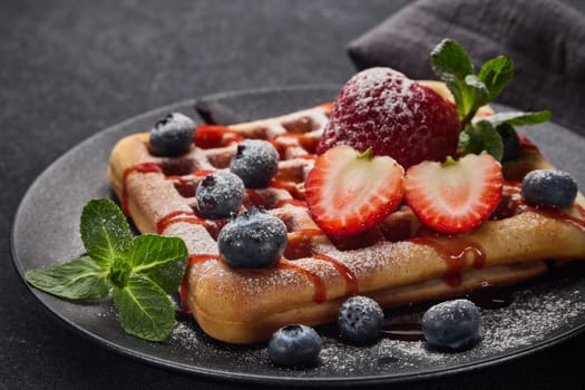 Delicious waffles with  powdered sugar and berries on ceramic black plate on grey background. Sweet meal. Dessert. Serving food