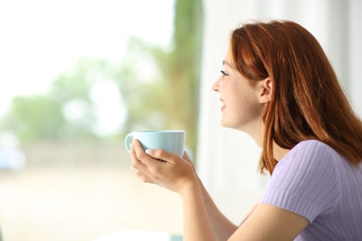 Happy woman drinking coffee looking through a window