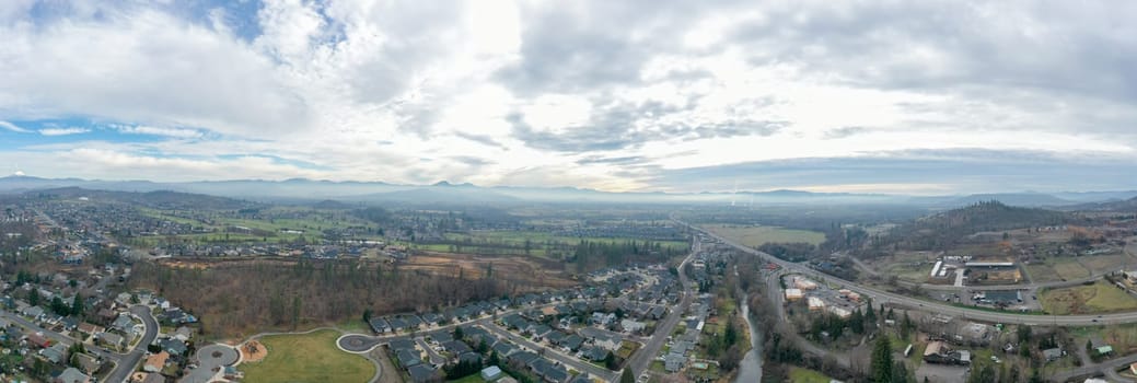 Wide-angle of peaceful small town under cloudy sky and with mountains as background. Aerial view of slightly cloudy blue skyline above the suburbs. Rural town landscape