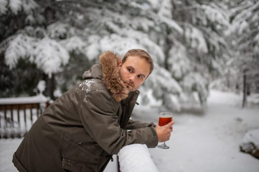 Man holding glass goblet leaning on snow-covered handrail with a snowy forest as background. Thoughtful man resting on wooden balcony and drinking. Relaxing winter holiday