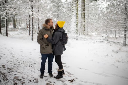Young couple smiling serenely and looking at each other in beautiful snowy forest. Lovable wife and husband surrounded by frozen trees. Lovely couples in winter holiday