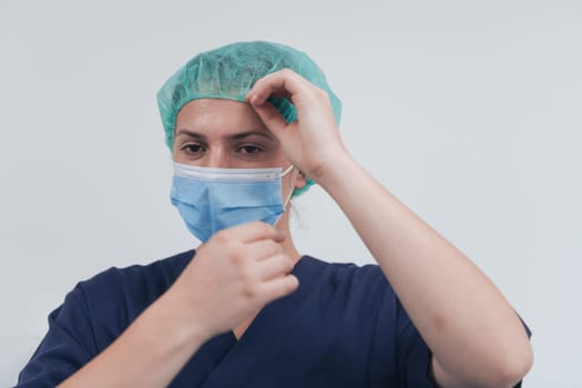 Close up of tired female doctor or scientist with a medical mask and surgical cap over grey background. Authentic real medical people hero in white in coronavirus pandemic is adjusting mask.