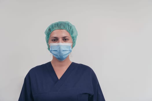 Close up of tired female doctor or scientist with a medical mask and surgical cap over grey background. Authentic real medical people hero in white in coronavirus pandemic is adjusting mask.