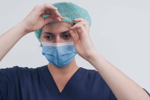 Close up of tired female doctor or scientist with a medical mask and surgical cap over grey background. Authentic real medical people hero in white in coronavirus pandemic is adjusting mask.