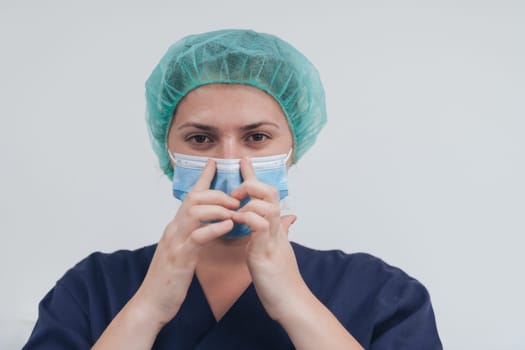 Close up of tired female doctor or scientist with a medical mask and surgical cap over grey background. Authentic real medical people hero in white in coronavirus pandemic is adjusting mask.