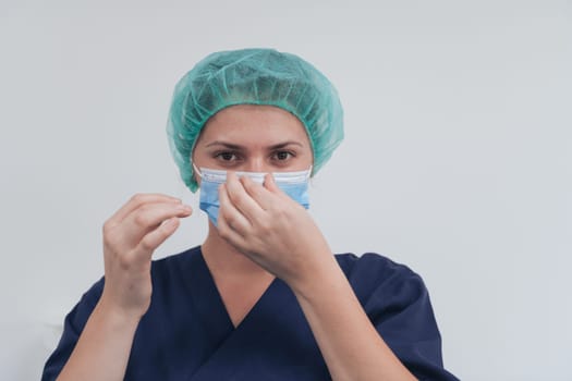 Close up of tired female doctor or scientist with a medical mask and surgical cap over grey background. Authentic real medical people hero in white in coronavirus pandemic is adjusting mask.