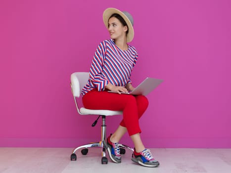 Portrait of young woman sitting on the chair and holding laptop on the lap isolated on pink background. Female model presenting fashion and technologz concept