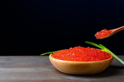 Red caviar in a wooden cup on a black background with a spoon and green onions. A large pile of bright caviar. Fresh delicious caviar. A place for advertising, logo, label, layout, mockup. Layout, template.