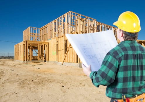 Contractor with Plans and Hard Hat Looking At New House Framing at Construction Site.
