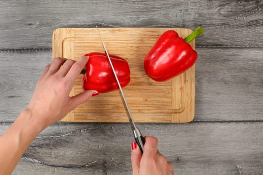 Top view, young woman hands holding knife cutting red bell pepper in half with other one placed on old chopping board.