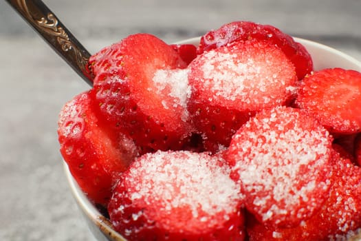Closeup photo of strawberries cut into circles, sprinkled with crystal sugar, in small bowl with silver spoon, ready to eat.