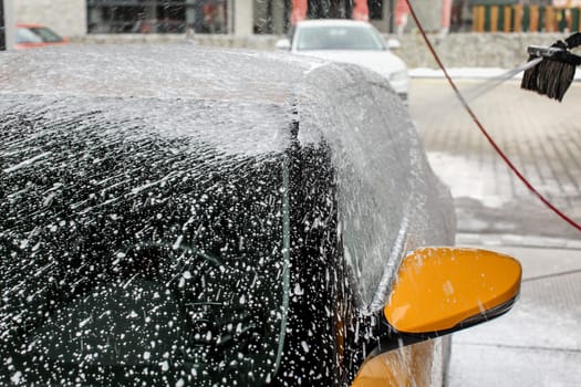 Yellow car almost completely covered in shampoo foam, with more spraying, when washed in carwash.