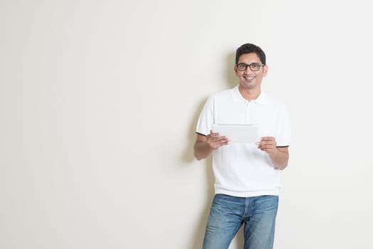 Portrait of handsome Indian guy using tablet pc, standing on plain background with shadow, copy space at side.