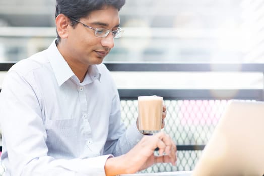 Asian Indian businessman using laptop computer while drinking a cup hot milk tea, outdoor cafe.