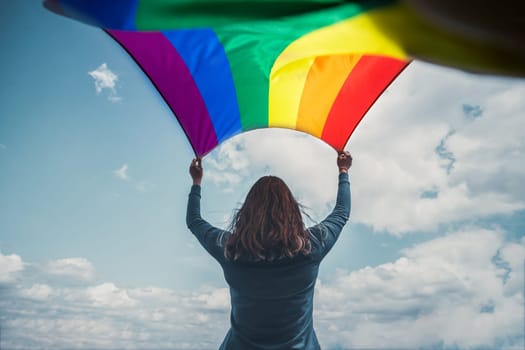 Woman holding a Gay Rainbow Flag over blue summer sky. Bisexual,gay, lesbian, transsexual symbol. Happiness, freedom and love concept for same sex couples