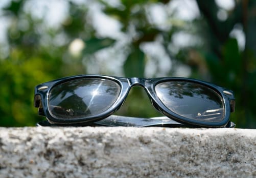 Black sunglasses with summer sunlight reflection placed on wall of a building with lens flare. Close up. Blurry defocused background. UV protection from ultraviolet light and sunburn heat concept.