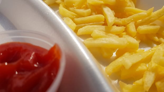 A close-up of golden orange yellow deep fries with tomato ketchup served in a foam container. Fast food takeaway. Unwanted fresh food