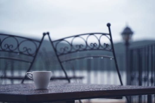 closeup detail of white small espresso coffee cup on wooden table with rain drops on rainy outdoor restaurant terrace