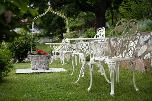 empty green wedding garden with red flowers in wicker basket and white Victorian metal decorative table and chairs